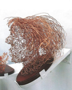 Resilience wire head with hair on wooden plinth attached to wall using sloping free standing plinth